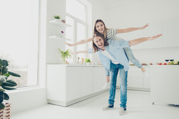 Portrait of cheerful lovely partners in casual outfits jeans making airplane holding open hands to sides, handsome man carrying on back lover in open space kitchen