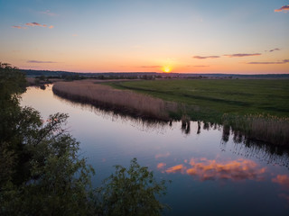 The calm surface of the river and the reflections of clouds, orange sunset, green fields and meadows in a quiet warm summer evening