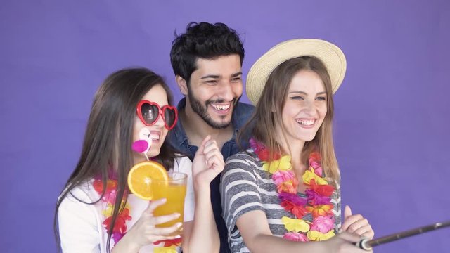 Friends taking selfie video with a selfie stick, two pretty long-haired girls in hawaiian wreaths and casual summer t-shirts smiling happily, handsome bearded man in denim blue shirt waving with the