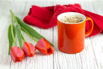 Three red tulips and a red mug of chocolate drink on a white wooden table.