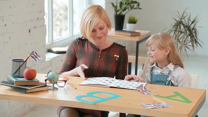 Student coming into the classroom for a lesson, smiling blond woman in checked red dress opens the book, little girl in denim outfit and white blouse waving 'hello', day shot in the modern classroom