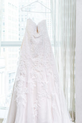 Long wedding dress hanging on hanger of window, with blinds, windowsill in urban New York City NY NYC high rise apartment condo building hotel