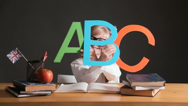 Primary pupil learning abc, wearing big black-framed glasses, having fun before the black wall as sitting at the desk with several books