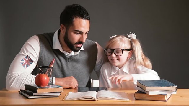 Intelligent male teacher in gray vest explaining new material to the primary student, bearded handsome man pointing at the text in book, blond funny girl listening to him cheerfully, indoor