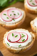 Bread slices with cream cheese, radish slices and chives on wooden board, photographed with natural light (Selective Focus, Focus on the front of the radish slices on the first bread)