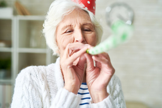 Head and shoulders portrait of happy senior woman blowing party horn and wearing birthday cap while enjoying holiday celebration at home, copy space