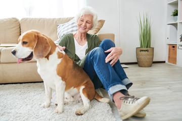 Full length portrait of happy  senior woman lovingly caressing pet dog sitting on floor in living room at home in modern apartment interior, copy space