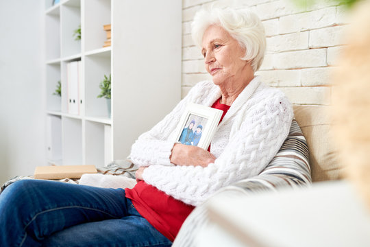 Side view portrait of devastated senior woman lovingly hugging framed photograph of her husband sitting at home alone in armchair by brick wall photo in frame by me .