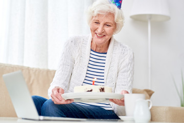 Portrait of happy senior woman showing cake to camera while celebrating birthday with her family via video chat