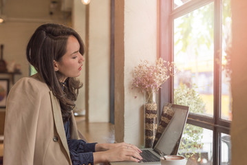 Young businesswoman sitting in coffee shop at table in front of laptop and drinking coffee. On background white brick wall and window. Girl shopping online, blogging, checking email.Online education.