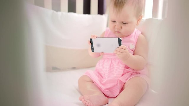 Little baby girl with phone in crib. Infant watching cartoon on mobile phone. Cute toddler looking smartphone in bed. Child with phone in cot