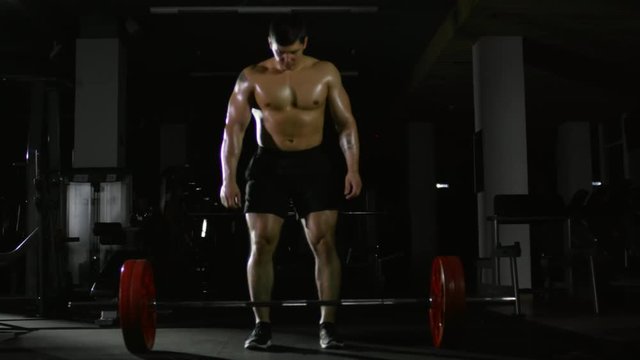 Full length of muscular shirtless man walking to heavy barbell in gym, picking it up from the floor and doing deadlift exercise