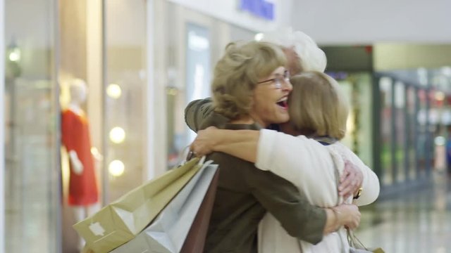 Group of three cheerful elderly women meeting each other in shopping mall, hugging and showing what they bought