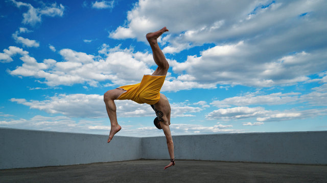 Tricking on street. Martial arts. Man makes back flip with arm support barefoot. Shooted from bottom foreshortening against sky.