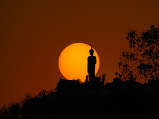 Silhouette of sunset behind buddha statue on hilltop.