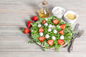 Delicious rucola salad with mozzarella, strawberries, olive oil and spices on a wooden background. Healthy foods