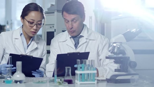 Tracking shot of two scientists discussing research project in laboratory and comparing data they have on clipboards