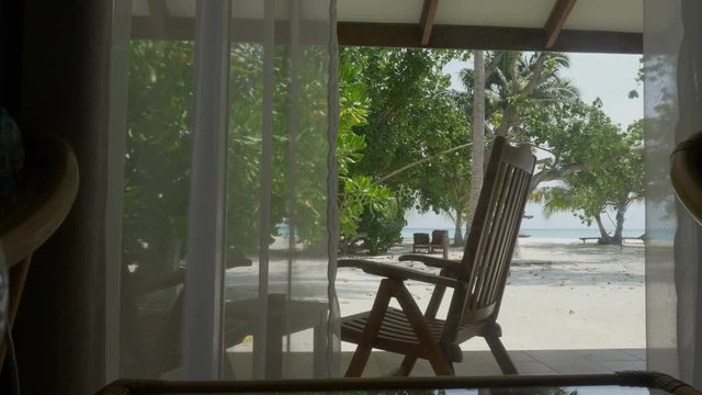 Window with curtain overlooking a beautiful beach with palm trees and ocean.