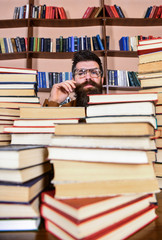 Man on thoughtful face between piles of books in library, bookshelves on background. Teacher or student with beard wears eyeglasses, sits at table with books, defocused. Scientific research concept.