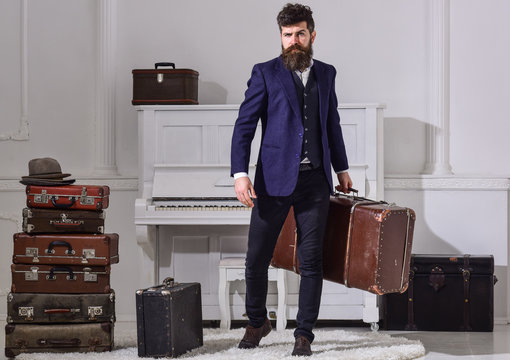 Macho stylish on strict face stands and carries big vintage suitcase. Man, traveller with beard and mustache with baggage, luxury white interior background. Baggage and travelling concept.