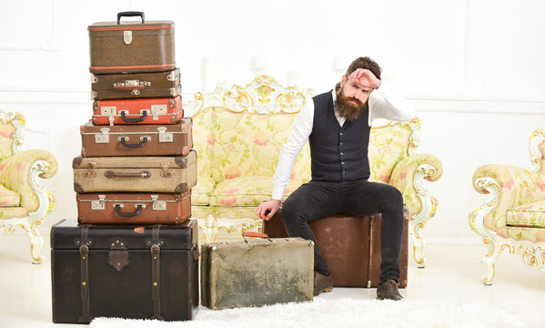 Macho elegant on strict face sits tired near pile of vintage suitcase. Man, butler with beard and mustache delivers luggage, luxury white interior background. Luggage and relocation concept.