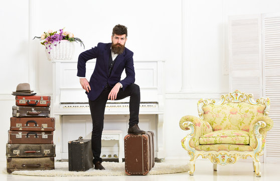Macho elegant on thoughtful face standing near pile of vintage suitcase. Man, traveller with beard and mustache with luggage, luxury white interior background. Luggage and travelling concept.