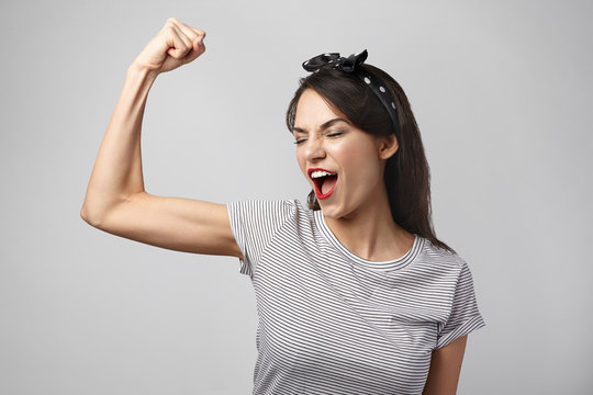 Picture of excited young brunette woman in casual clothes opning mouth widely while screaming, showing off muscles, tensing bicep, demonstrating power and energy. Sports, fitness and strength concept