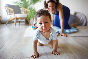 Horizontal shot of cute funny infant crawling along floor from his mother who is standing on her knees in background, trying to catch her baby son, smiling happily. Mom and child playing at home - obrazy, fototapety, plakaty