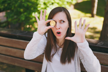 Close up fun emotional young female in light casual clothes. Woman sitting on bench holding bitcoin in front eye, coin of golden color in street outdoors. Online virtual future currency concept.