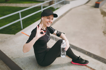 Young athletic smiling girl in black uniform, cap with headphones listening music, holding bottle with water, sitting on training, showing OK sign in city park outdoors. Fitness, healthy lifestyle.
