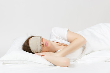 Calm young brunette woman lying in bed with sleep mask on white sheet, pillow, blanket on white background. Smiling beauty female spending time in room. Rest, relax, good mood concept. Copy space.