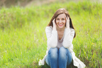 Young brunette smiling beautiful woman in light casual clothes keeping hands near face sitting on grass in sunny day on green field background. Beautiful landscape. Lifestyle, leisure concept.