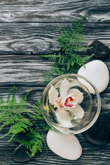 flat lay with spa and massage treatment arrangement with fern, pebbles and orchid flower in bowl on wooden tabletop