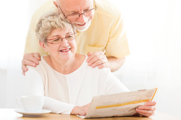 Smiling senior woman reading newspaper while husband hugging her in the morning