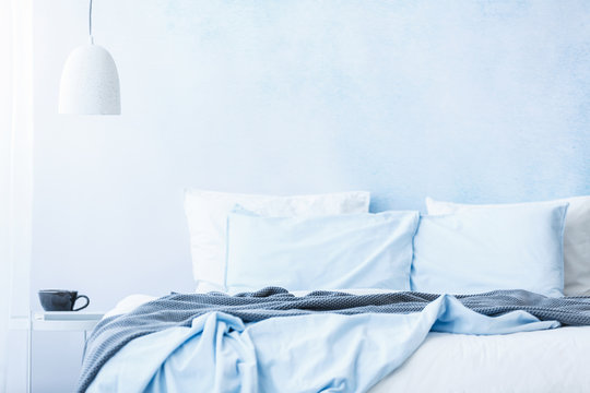 Blue Bedsheets And Pillows On Bed Next To A Table With A Cup Under White Lamp In Simple Bedroom Interior