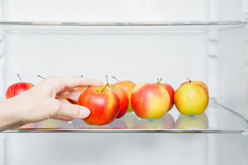 Woman's hand taking beautiful, fresh, colorful apple from fridge shelf in the kitchen. Healthy sweet food concept. New start for healthy nutrition, body slimming, weight loss. Cares about body.