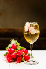 A photo of a light yellow cold summer aperitif alcoholic cocktail in a wine glass, full of ice cubes, garnished with radish. White wooden table.