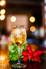 A photo of a light yellow cold summer aperitif alcoholic cocktail in a wine glass, full of ice cubes, garnished with radish. White wooden table.