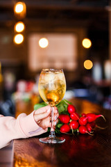 A photo of a light yellow cold summer aperitif alcoholic cocktail in a wine glass, full of ice cubes, garnished with radish. White wooden table.