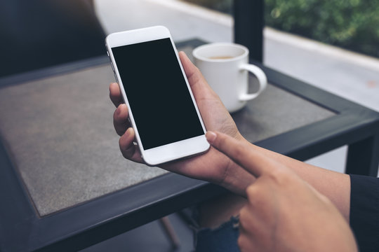 Mockup image of hands showing and pointing at a white mobile phone with blank black desktop screen in cafe