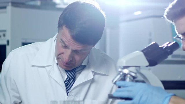 Medium shot of two male scientists working in laboratory, one using microscope and one writing down research information