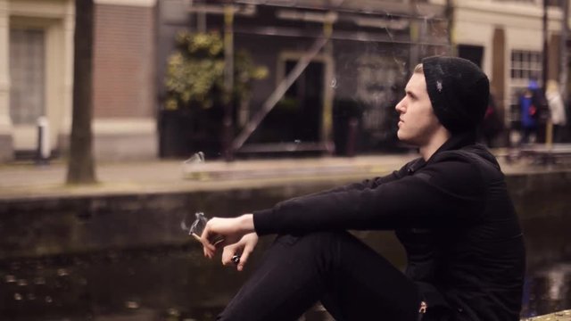 Man smoking marijuana in amsterdam. Boy smoking a marihuana joint on the street in Amsterdam. Young boy smoking weed on river background in slow-mo. 60-fp. Tourist try marijuana near coffee shop .