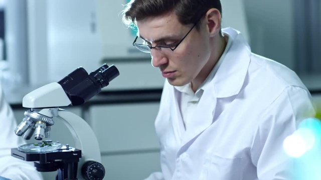 Medium shot of young man in lab coat, glasses and rubber gloves using microscope to study plant specimen when working in laboratory