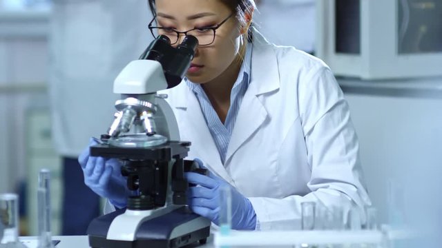 Tilt up shot of young Asian woman in lab coat, glasses and rubber gloves using microscope in laboratory, medium shot