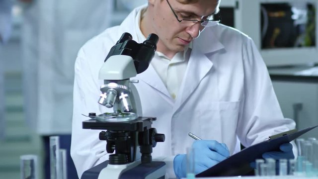 Medium shot of young chemist in glasses writing down research details on paper attached to clipboard when studying plant specimen under microscope