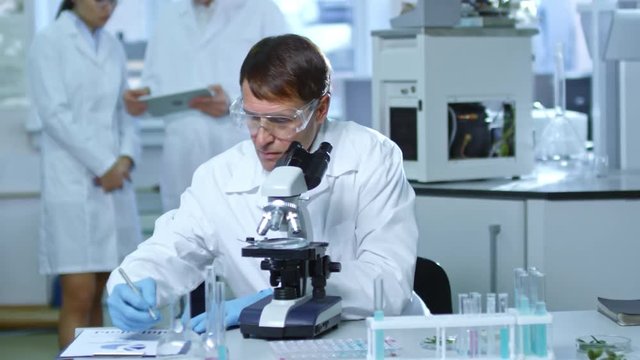 Mature man sitting at table in laboratory and studying samples under microscope, then writing down test results