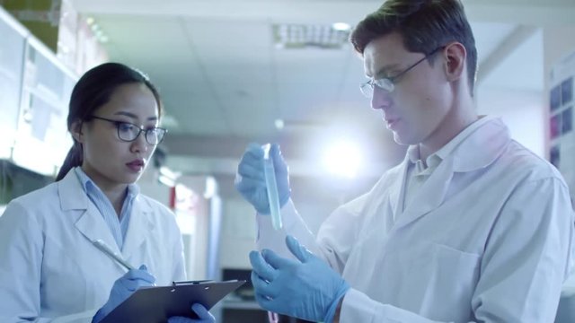 Medium shot of two scientists in lab coats, rubber gloves and glasses analyzing contents of test tube and writing down information on their research, tilt up
