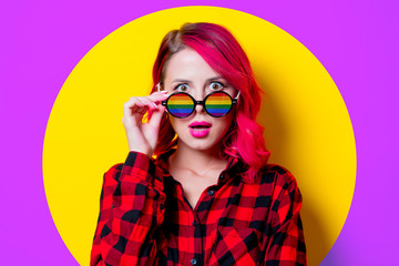 Young pink hair girl in red tartan shirt and rainbow sunglasses. Portrait isolated on yellow background