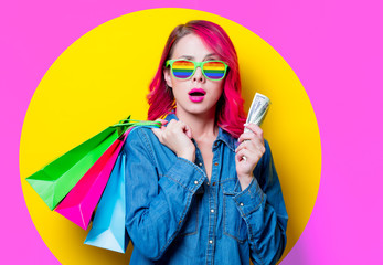 Young pink hair girl in blue shirt and rainbow glasses holding a colored shopping bags with money. Portrait isolated on yellow background