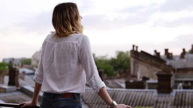 View from behind of beautiful woman stands on the balcony or terrace in apartment at day time.Young lady enjoying the view on the balcony of her hotel room. Small buildings and houses view